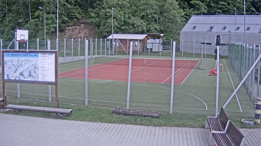 An outdoor tennis court and basketball court are enclosed by a fence, with benches and a building visible in the background on an overcast day.