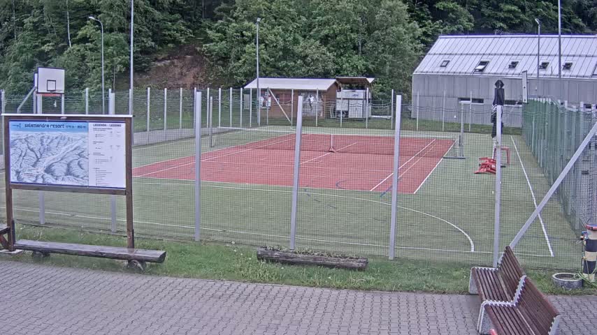 An outdoor multi-purpose court with a red surface and green surrounding area is enclosed by a chain-link fence, under an overcast sky.