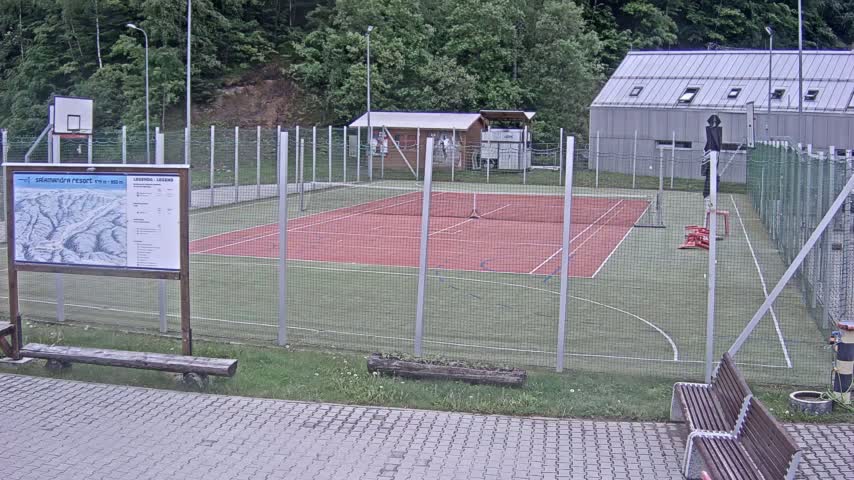 An outdoor multi-use court with a red surface and green perimeter, enclosed by a chain-link fence, is situated near a building and some trees on an overcast day.