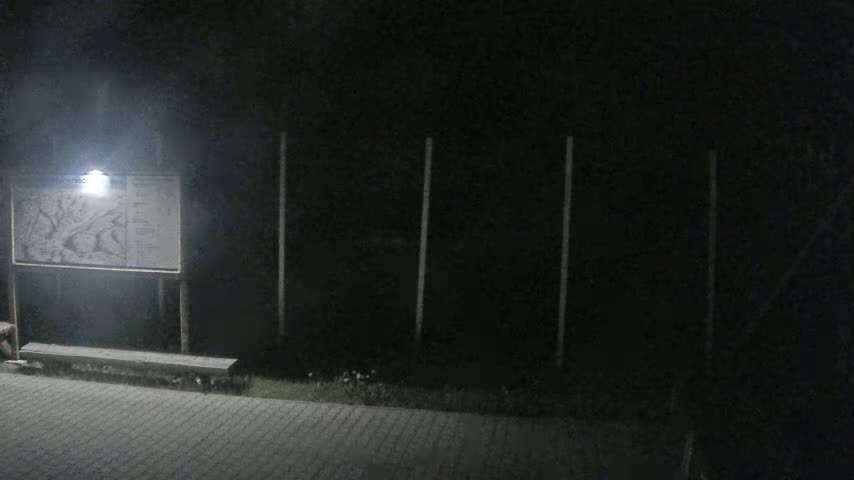 A dimly lit nighttime scene shows a sign with a map, a bench, and a dark fence.