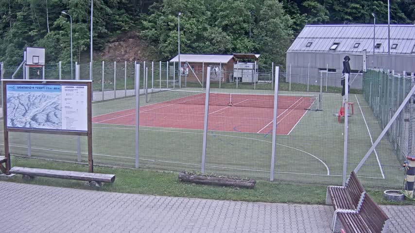 An outdoor multi-purpose court with a red surface, enclosed by a chain-link fence, sits next to a small building under an overcast sky.
