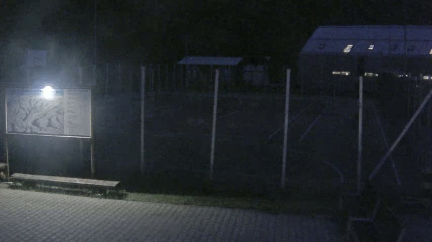 A nighttime scene shows a lit map display and bench next to a chain-link fence surrounding a dark, empty playing field and building.