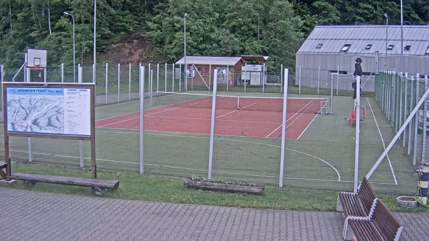 An outdoor tennis and basketball court with a small building and surrounding green space is visible on an overcast day.