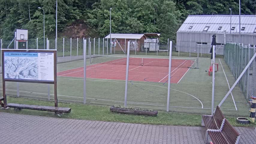 An outdoor multi-purpose court with a red tennis court surface and a green basketball court surface is enclosed by a chain link fence, under an overcast sky.
