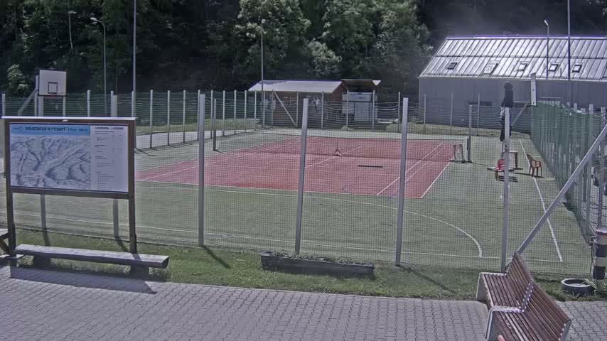 A fenced-in outdoor tennis and basketball court with a small building and benches sits amidst a wooded area on a sunny day.