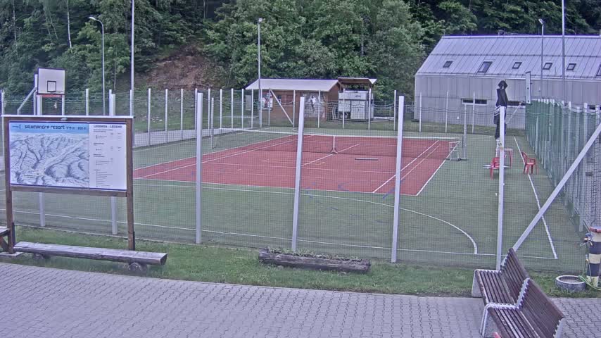 An outdoor multi-purpose court with a red surface, surrounded by a chain-link fence, sits near a small building and a wooded area on an overcast day.