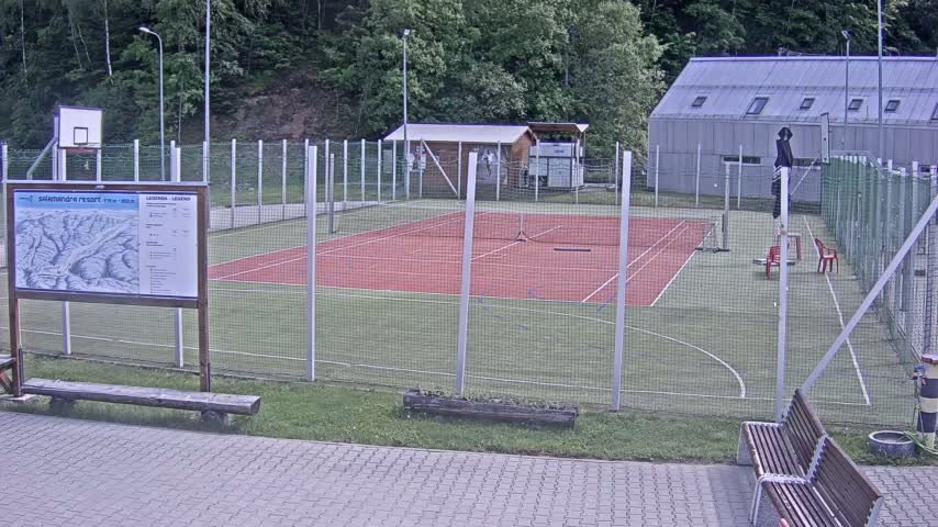 An outdoor multi-use sports court with a red surface, surrounded by a chain-link fence, is situated next to a building under overcast skies.
