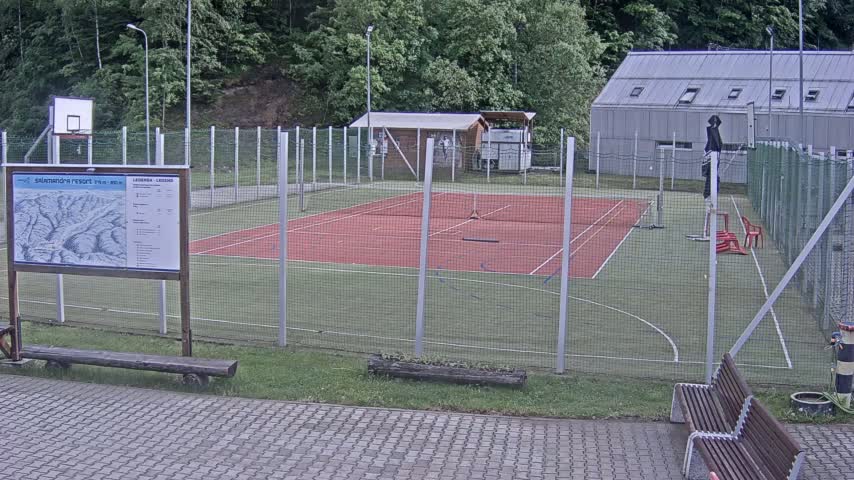 An outdoor tennis court and basketball court are enclosed by a chain-link fence, with a small building and some trees in the background under an overcast sky.