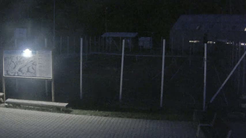A lit sign with a map is next to a bench in front of a fenced-in area with a building at night.
