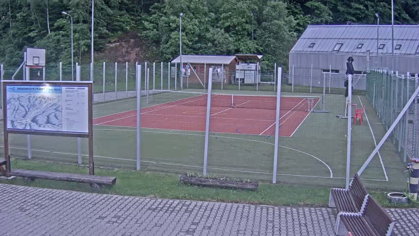 An outdoor multi-use court with a red surface, enclosed by a chain-link fence, sits adjacent to a small building under an overcast sky.