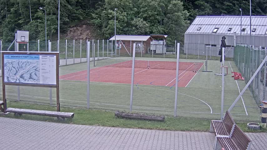 An outdoor multi-use sports court with a red surface, surrounded by a chain-link fence, sits beside a small building on a partly cloudy day.