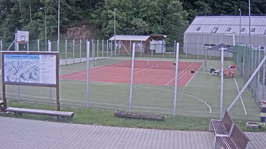 An outdoor tennis court with a basketball hoop, surrounded by a chain-link fence, sits next to a small building under an overcast sky.