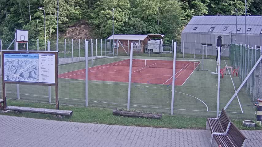 An outdoor multi-use sports court with a red surface and green surrounding area, enclosed by a chain-link fence, is visible under overcast conditions.