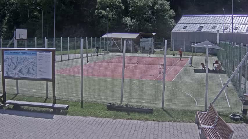 A fenced-in tennis court with a red surface and green perimeter, a basketball hoop, a small building, and several people are visible on a sunny day.