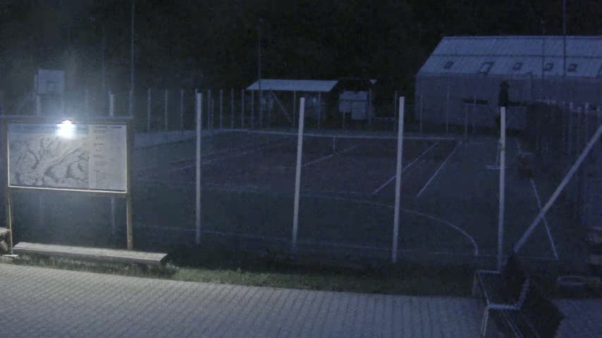 A nighttime view of an outdoor sports court surrounded by a fence, with a map and a bench nearby.