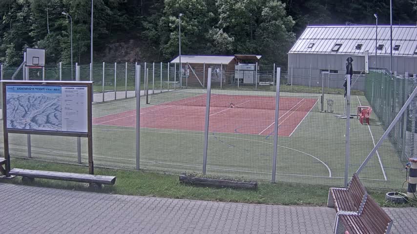 An outdoor tennis court and basketball court are enclosed by a chain-link fence, with a small building and wooded area visible in the background on a cloudy day.