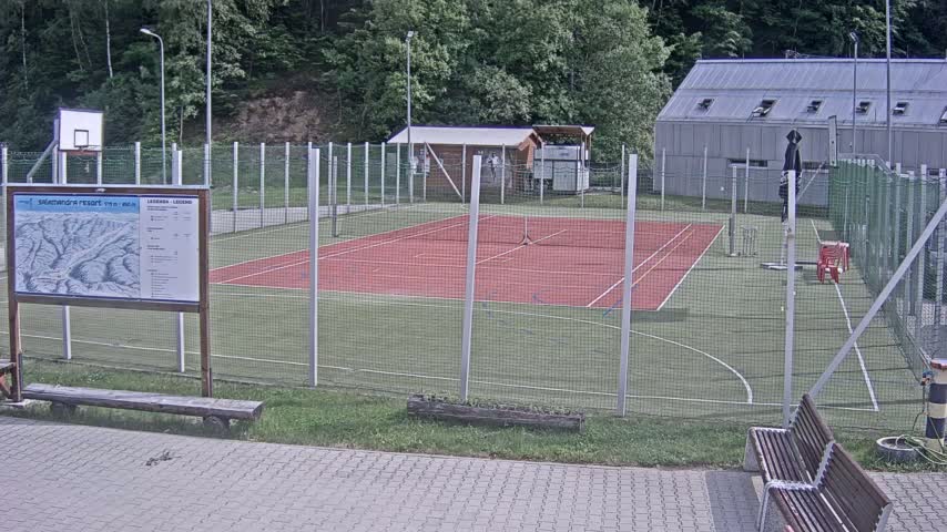 An outdoor multi-purpose court with a red surface, surrounded by a chain-link fence, is situated in a wooded area on an overcast day.