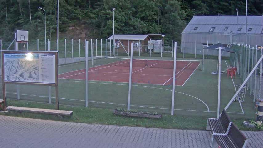 An outdoor tennis and basketball court with a small building and benches sits behind a chain-link fence on a partly cloudy evening.