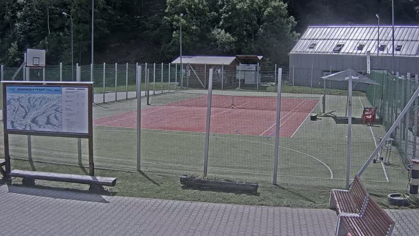 An outdoor tennis court and basketball court are enclosed by a chain link fence, with a building and a few benches nearby on a sunny day.