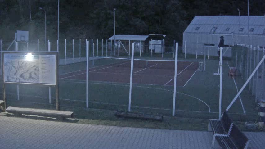 An outdoor tennis and basketball court, enclosed by fencing, is dimly lit at night.
