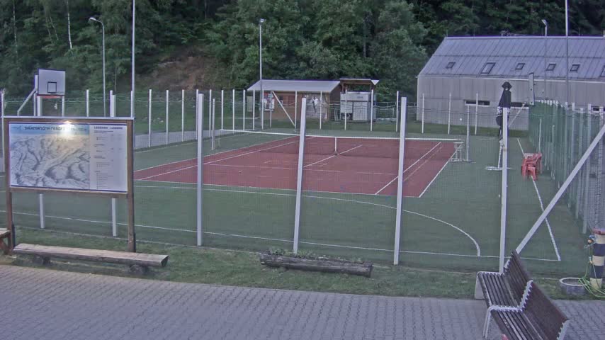 An outdoor multi-use court with a red tennis/basketball court surrounded by a fence, a small building, and benches.