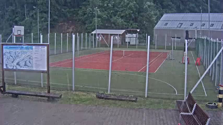 An outdoor multi-purpose sports court with a basketball hoop and tennis lines is surrounded by a chain-link fence in rainy weather.