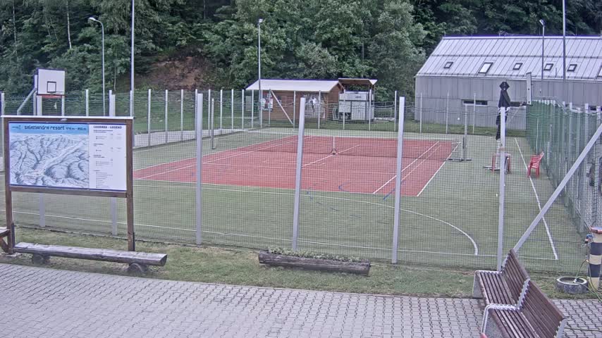 An outdoor multi-use sports court with a red surface, enclosed by a chain-link fence, sits near a small building and basketball hoop, all under an overcast sky.