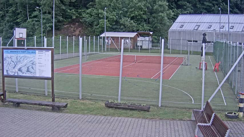 A fenced-in outdoor sports court with a red tennis court and green artificial turf, surrounded by trees and a small building, on an overcast day.