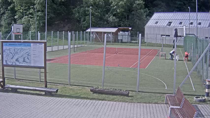 A red multi-purpose court enclosed by a chain-link fence is situated outdoors on a sunny day, next to a building and a basketball hoop.