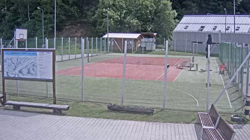An outdoor tennis and basketball court enclosed by a chain-link fence on a sunny day, with a small building and trees in the background.