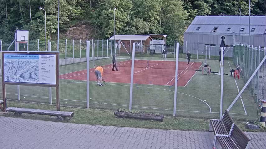 Several people play tennis on an outdoor court surrounded by a chain-link fence, under a partly cloudy sky.