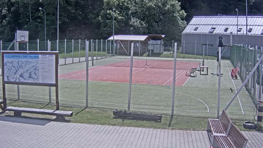 An outdoor tennis court with a basketball hoop and surrounding fencing is visible on a sunny day.