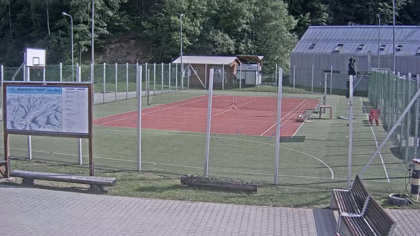 An outdoor multi-purpose sports court with a red surface, surrounded by a chain-link fence, on a sunny day.