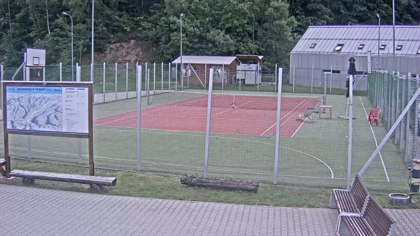 An outdoor tennis court with a basketball hoop is enclosed by a chain-link fence, and is situated next to a building on a mostly sunny day.
