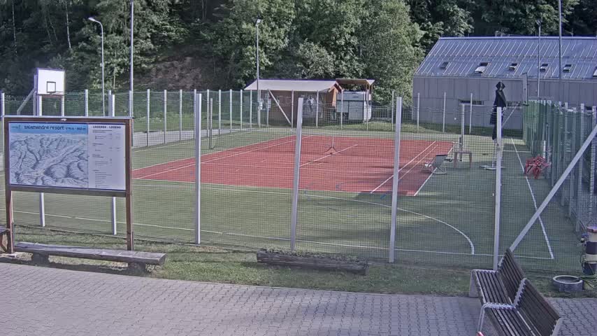 An outdoor tennis court with a basketball hoop and surrounding fence is visible on a partly sunny day.