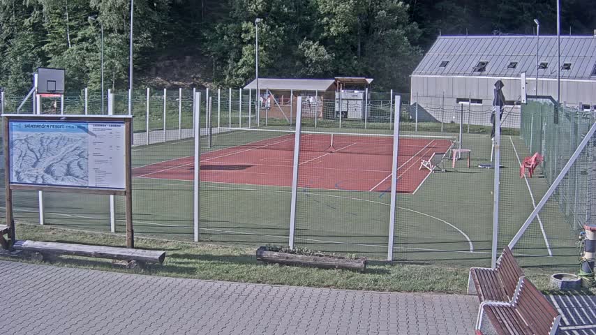 An outdoor tennis and basketball court complex is surrounded by a chain-link fence on a sunny day.