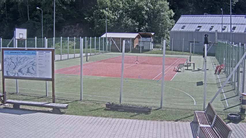 A red tennis court enclosed by a chain-link fence is surrounded by green grass, a building, and trees on a sunny day.