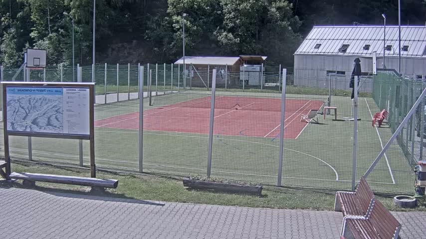 An outdoor tennis court and basketball court are enclosed by a chain-link fence, with a building and trees visible in the background on a sunny day.