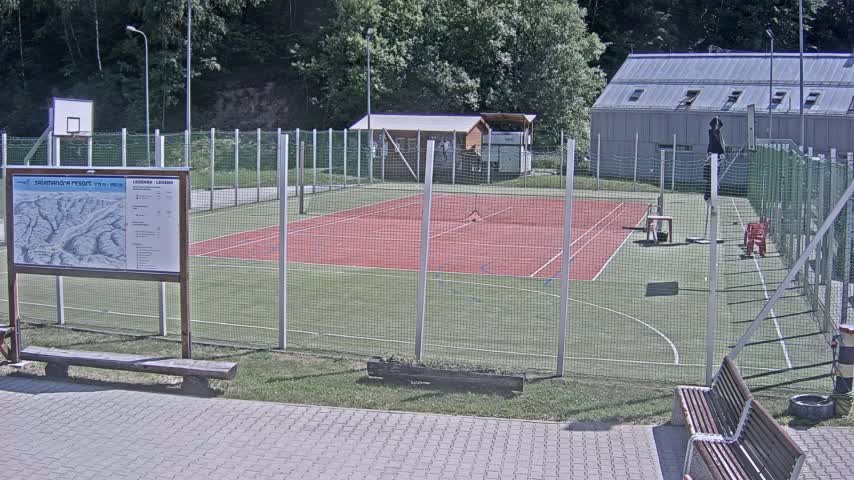 An outdoor tennis court with a basketball hoop and surrounding fenced area, on a sunny day, is shown next to a building.
