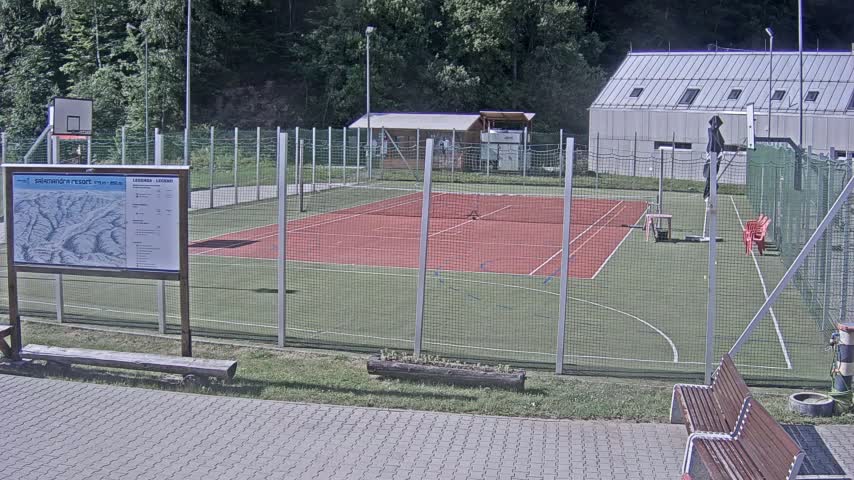 An outdoor multi-use sports court with a red surface, surrounded by a chain-link fence, sits next to a building and a basketball hoop.