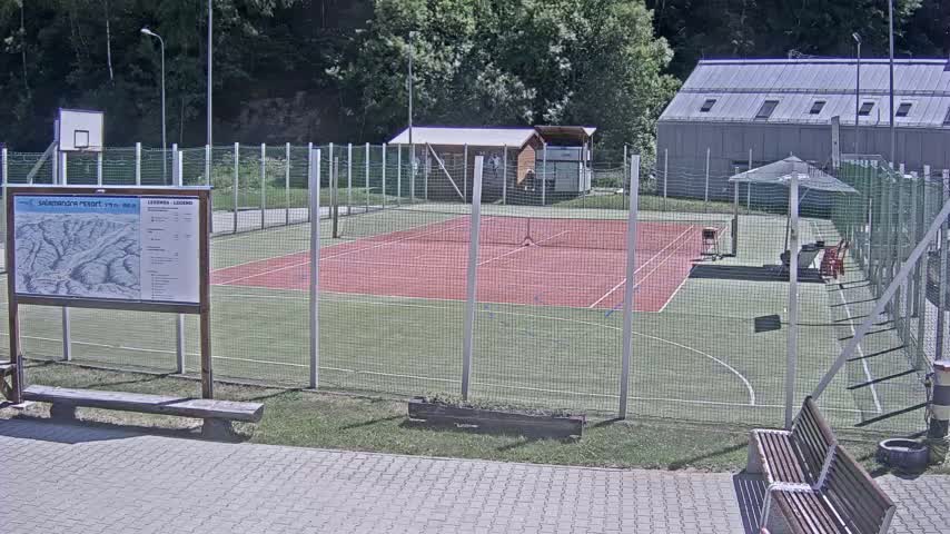 An outdoor tennis court, enclosed by a chain-link fence, sits next to a basketball court on a sunny day.
