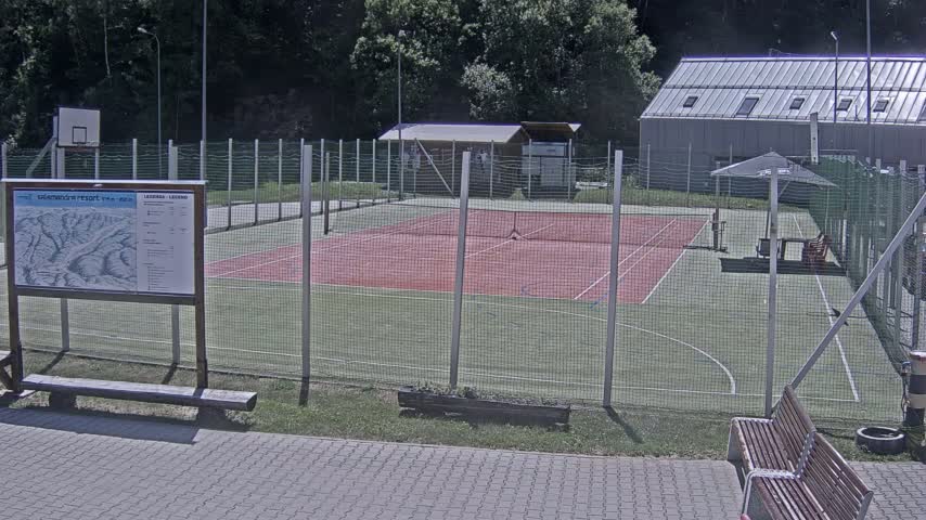An outdoor tennis/multi-purpose court with a basketball hoop, enclosed by a chain-link fence, is visible on a sunny day.