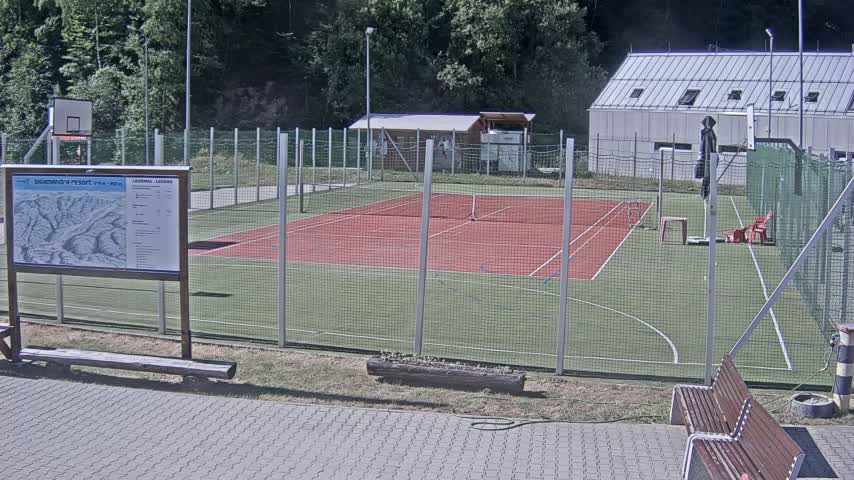 A red tennis court enclosed by a chain-link fence, with a basketball hoop and benches visible, sits outdoors on a sunny day.