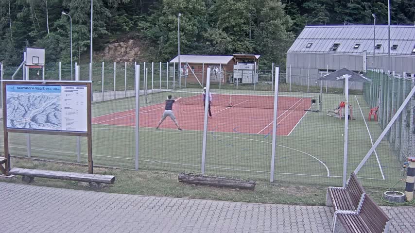 Two people play tennis on an outdoor court surrounded by a chain-link fence on a partly cloudy day.