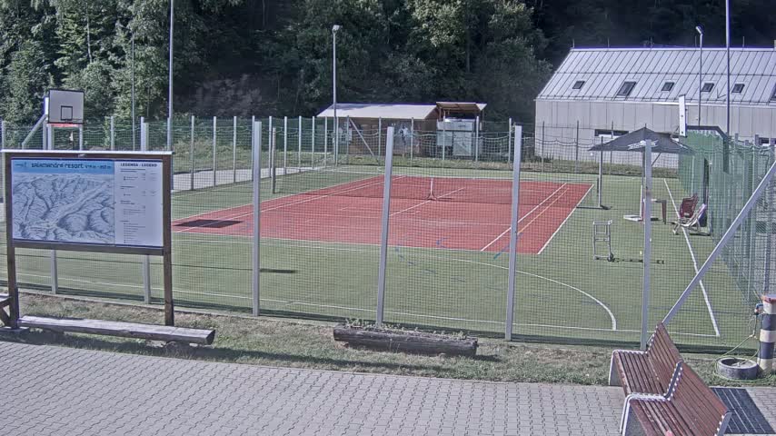 An outdoor tennis court and basketball court are enclosed by a chain link fence on a sunny day, with a building and trees in the background.