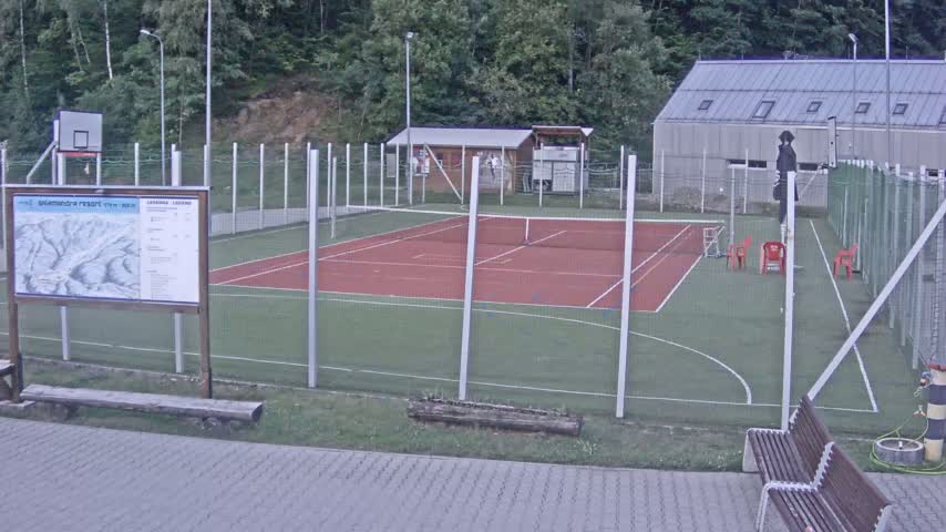 An outdoor tennis court and basketball court are enclosed by a chain link fence on a sunny day, with a building and trees in the background.