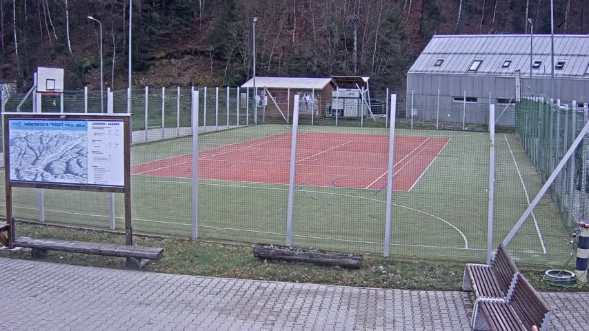 A tennis court with two people on it, one appearing to be maintaining it and the other playing, is surrounded by a fence, with a basketball court and a building in the background on a sunny day.
