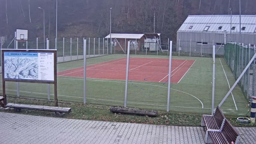 An outdoor multi-purpose sports court with a red playing surface and green outfield is visible behind a fence, flanked by buildings and bare trees under a dull, overcast sky, with paved areas and benches in the foreground.