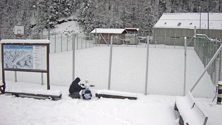 Under overcast skies on a snowy winter day, two figures are building a snowman in a fenced outdoor area, with snow-covered benches, an information board, and buildings, all backed by a dense forest of snow-laden trees.