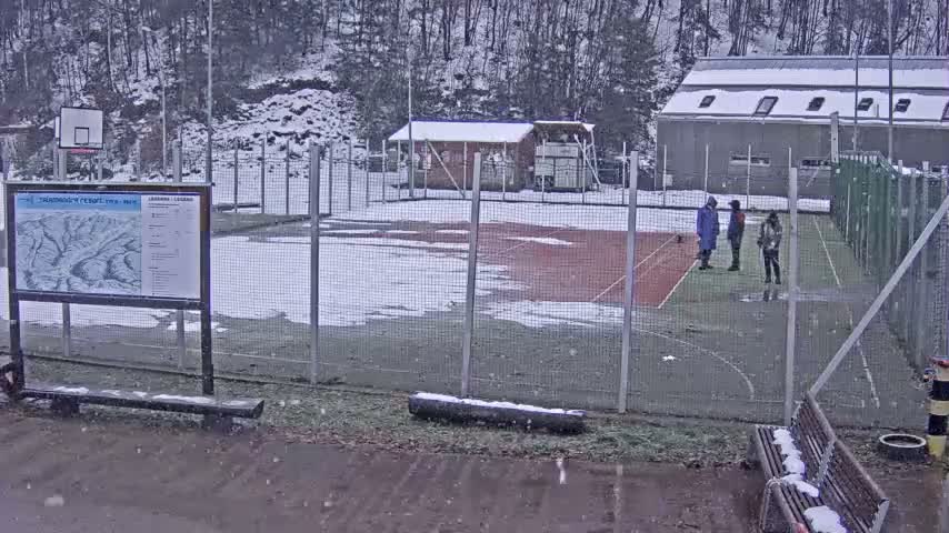 Light snow falls over a fenced outdoor sports court and surrounding area, where snow covers the ground, benches, and roofs of distant buildings, while three individuals stand on the partially snow-dusted court surface.